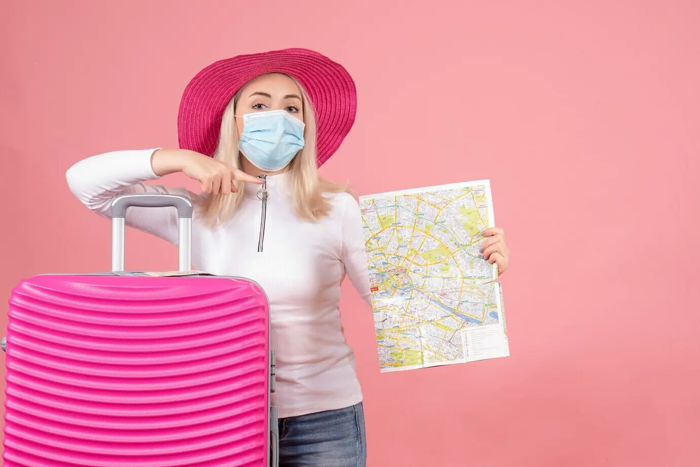 Вид спереди: симпатичная молодая девушка стоит рядом с чемоданом, держит карту и указывает на что-то.