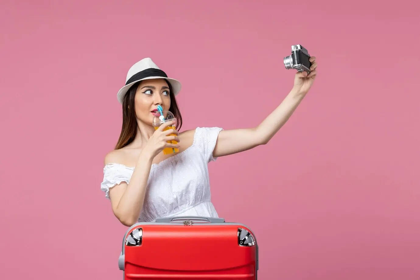 Вид спереди: молодая женщина фотографирует на отдыхе на фоне розовой стены; летние цвета, отпускная поездка, жара, путешествие.